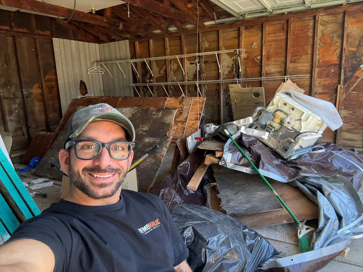 Garage packed with stacked demolition panels, black trash bags, a stripped appliance shell and a broom, with a crew member in frame
