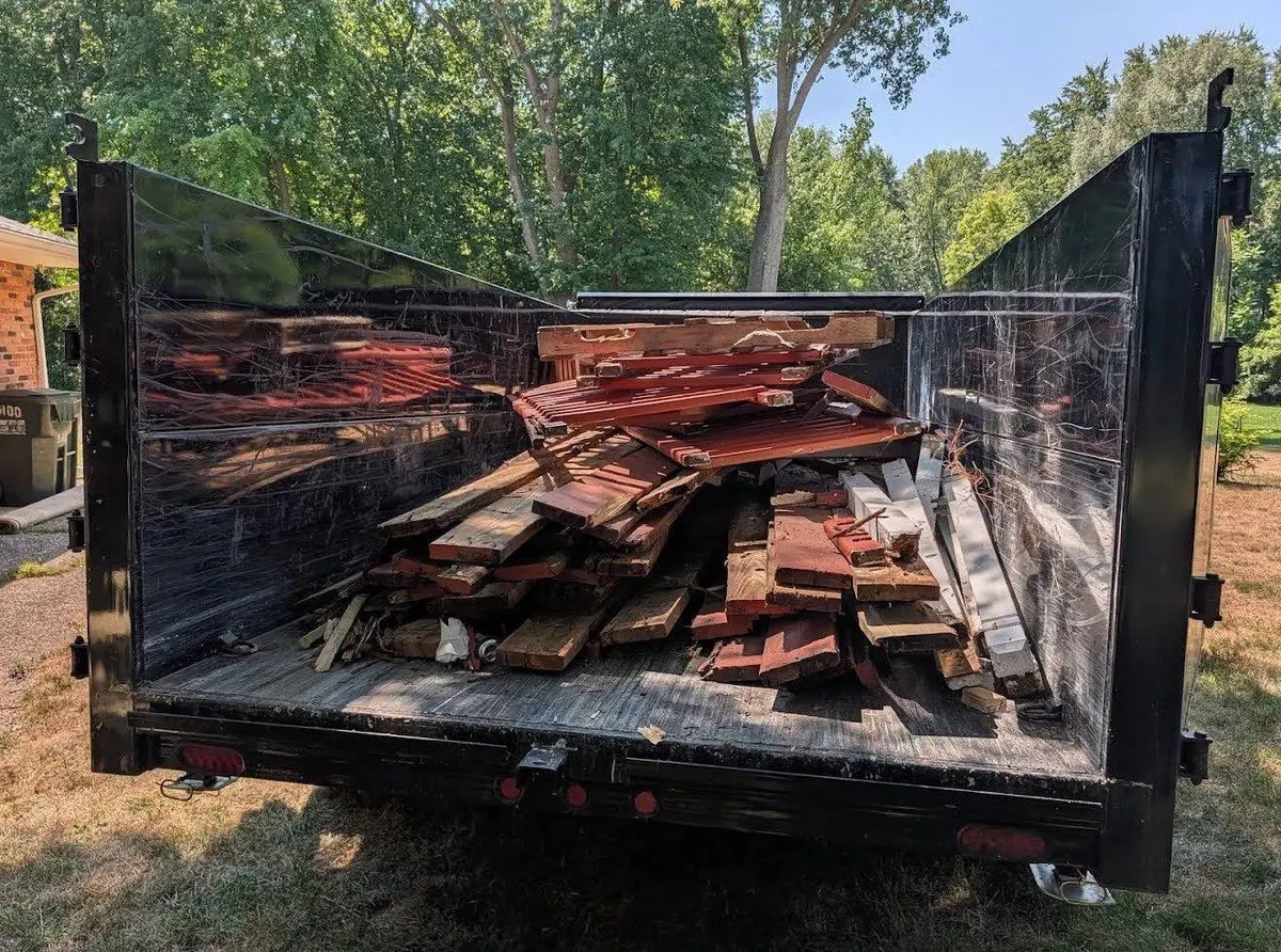Black trailer loaded with stacked red-toned lumber boards and wrapped material, viewed from behind with trees in the background