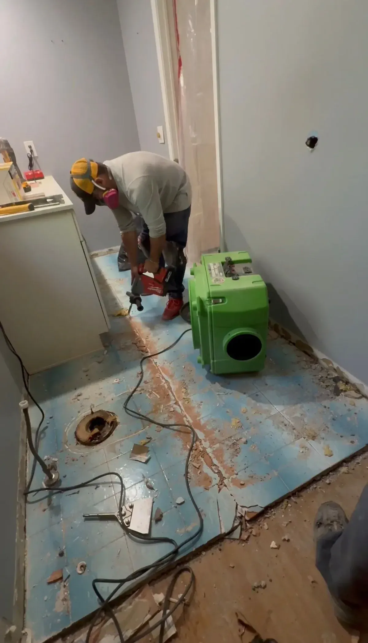Demohaulic crew member in a yellow hard hat and pink half-mask respirator cutting blue bathroom floor tile with a red demolition hammer beside a running green HEPA air scrubber; blue floor protection, a waste pipe stub, broken tile debris, and a white vanity cabinet are visible