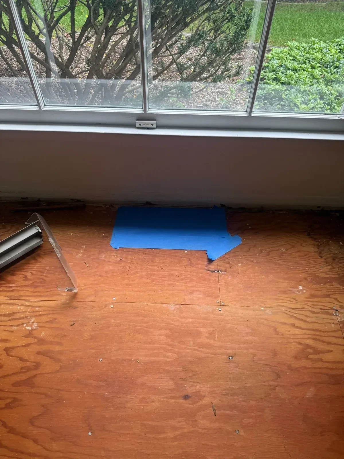 Plywood floor by a kitchen window with blue painter’s tape covering a floor register and clear plastic protection visible at the edge