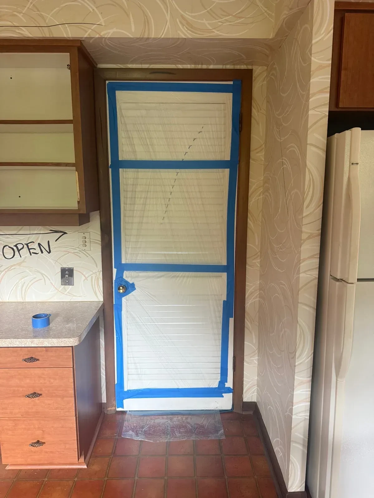 Kitchen doorway sealed with a clear plastic ZIPWALL zipper door and blue painter’s tape, with cabinets and refrigerator still in place