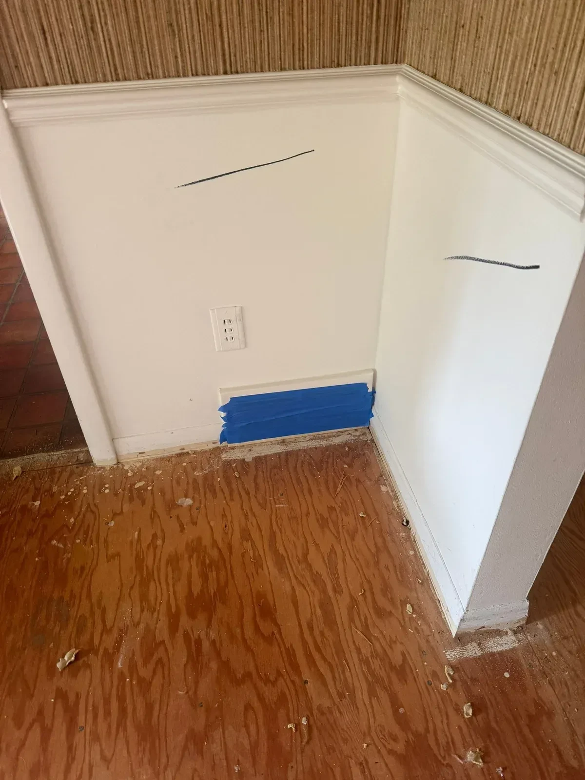 Kitchen wall corner with blue painter’s tape covering a low register, marked wall lines and a plywood floor during demolition