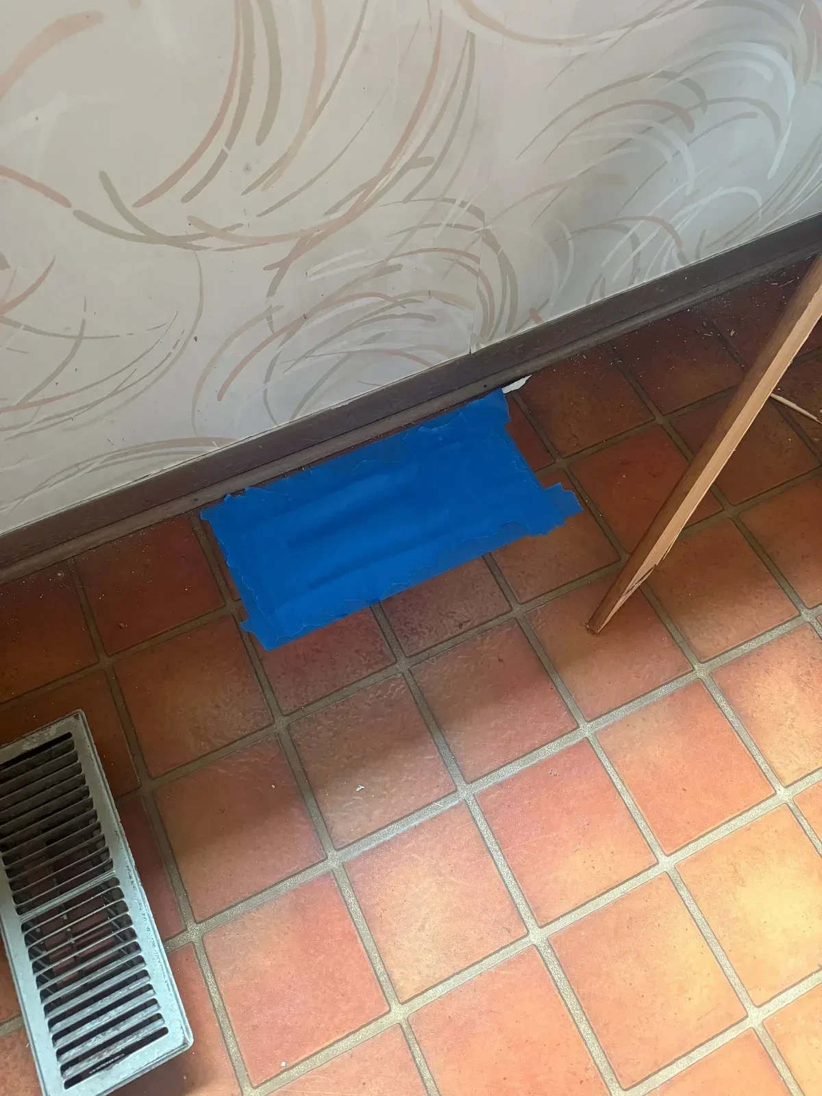 Kitchen wall with swirled wallpaper and a blue-taped wall register above the red-brown tile floor during demolition