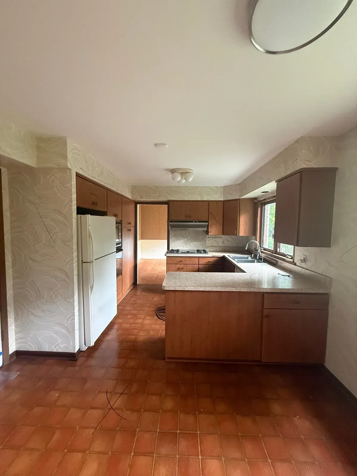 Intact galley kitchen before demolition with wood cabinets, white refrigerator, wraparound counter, tile floor and patterned wallpaper