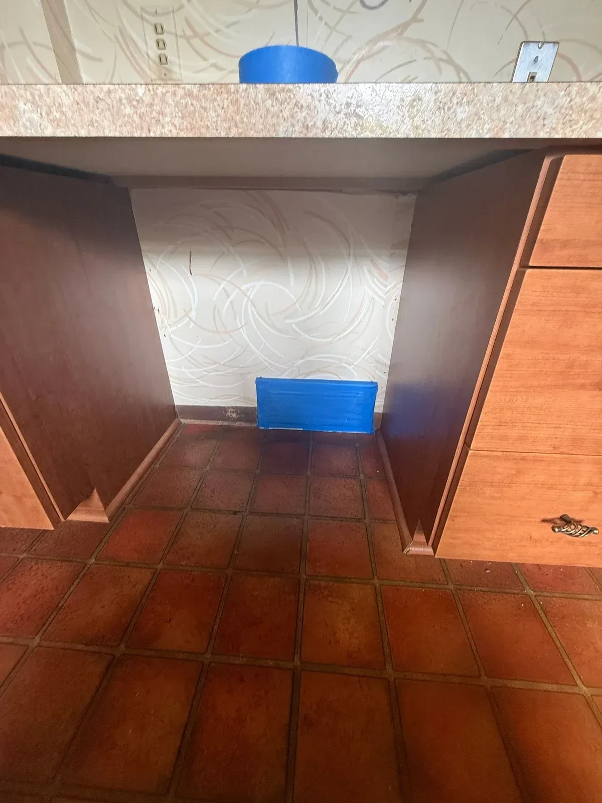 Red-brown kitchen tile floor between wood-toned cabinets with blue painter’s tape covering a wall register