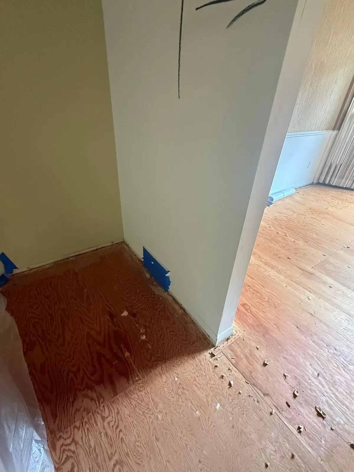 Kitchen floor stripped to plywood subfloor with blue painter’s tape over a wall register and demolition debris near the doorway