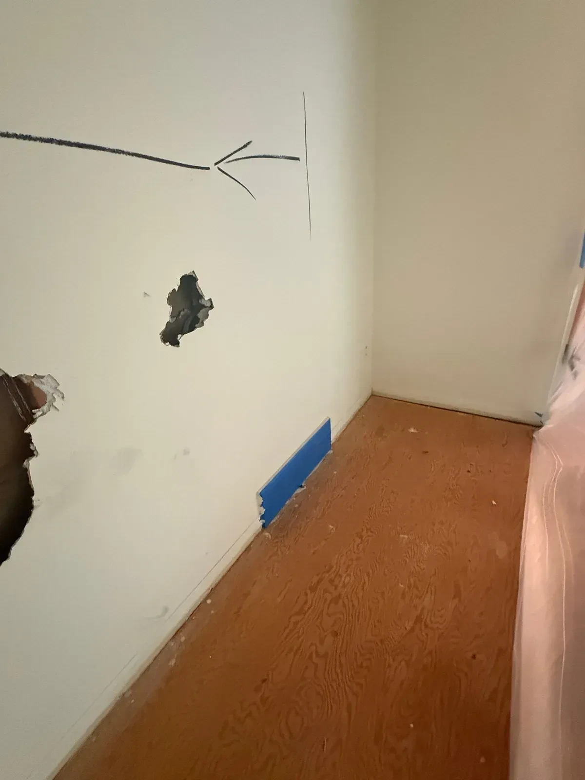 Stripped plywood subfloor with plastic drop sheeting, blue tape over a wall register and painted cut marks on the wall