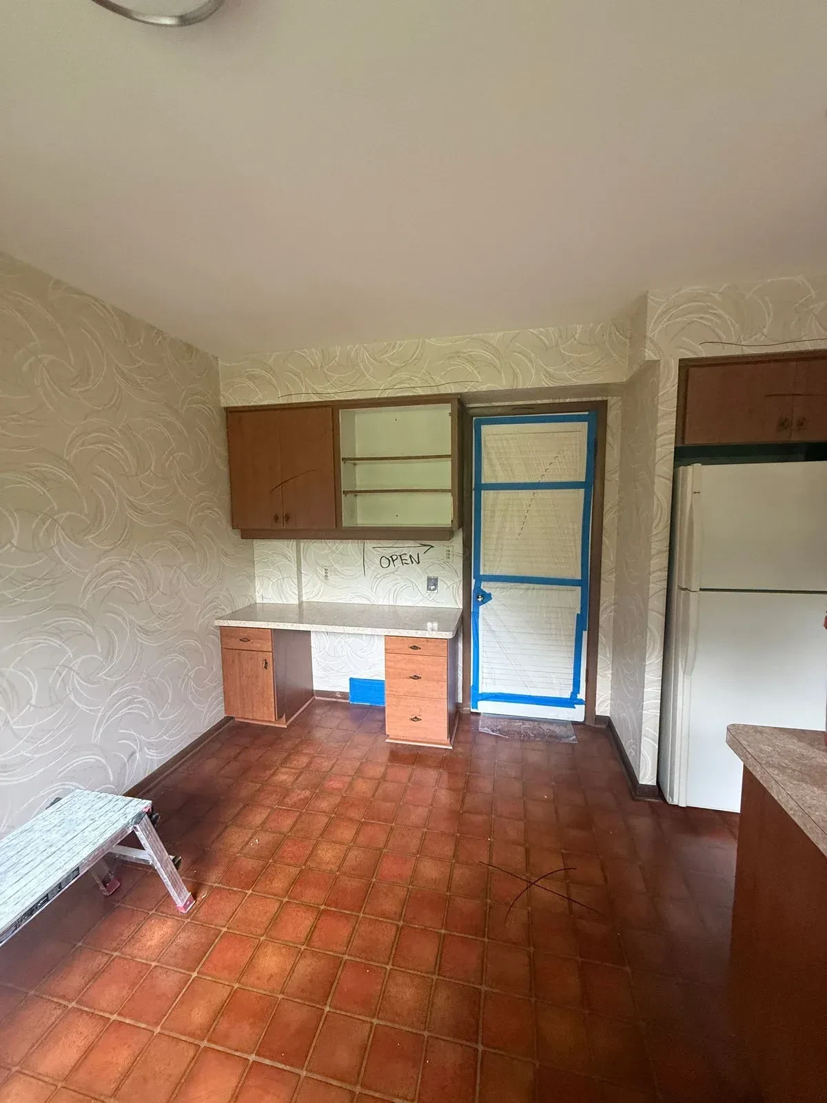 Galley kitchen before demolition with wood-toned cabinets, built-in desk and hutch, white refrigerator, red-brown tile floor and swirled-pattern wallpaper