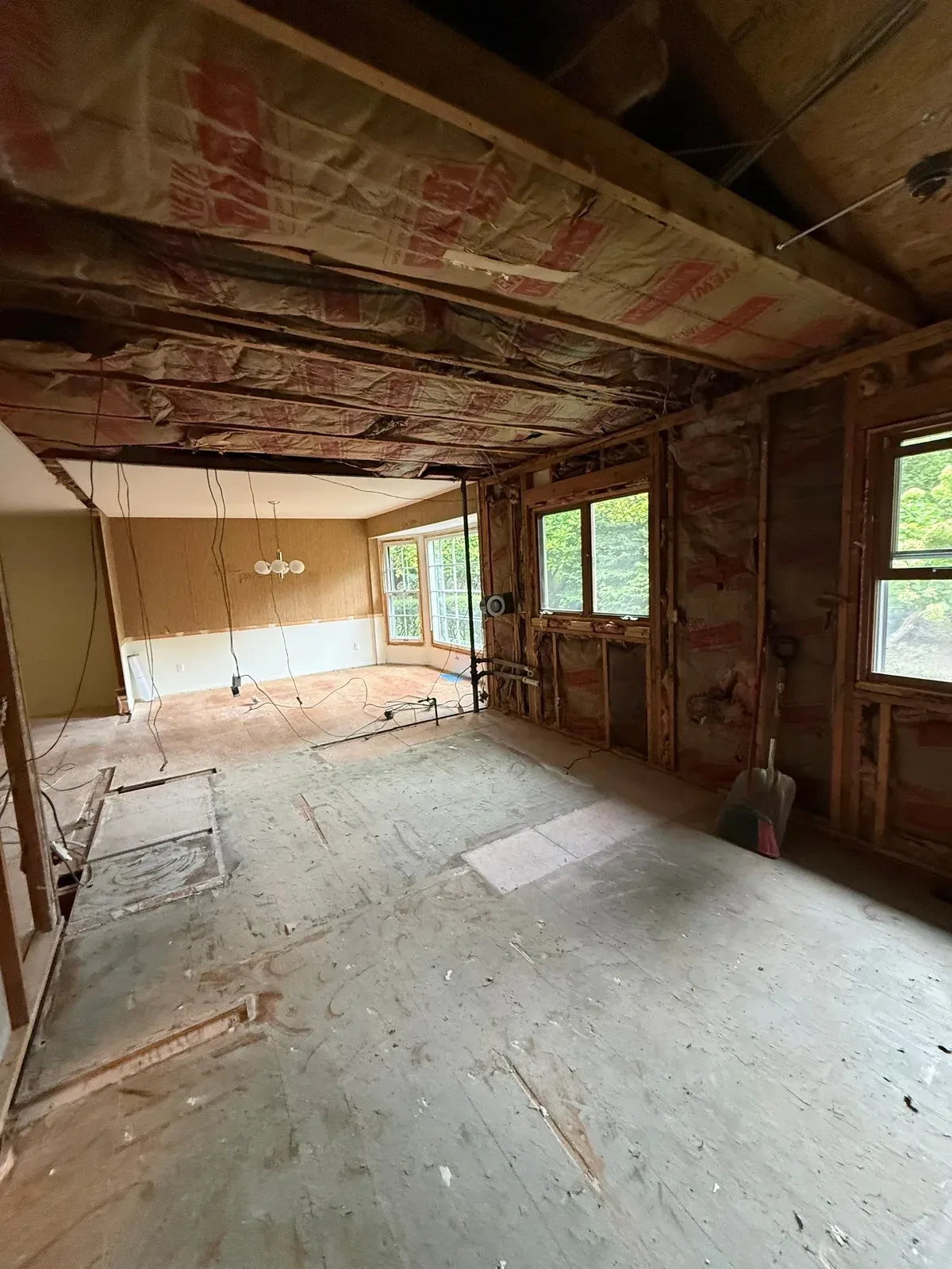 Opened kitchen and living area with exposed wall framing, ceiling insulation, stripped floor and demolition equipment near the windows