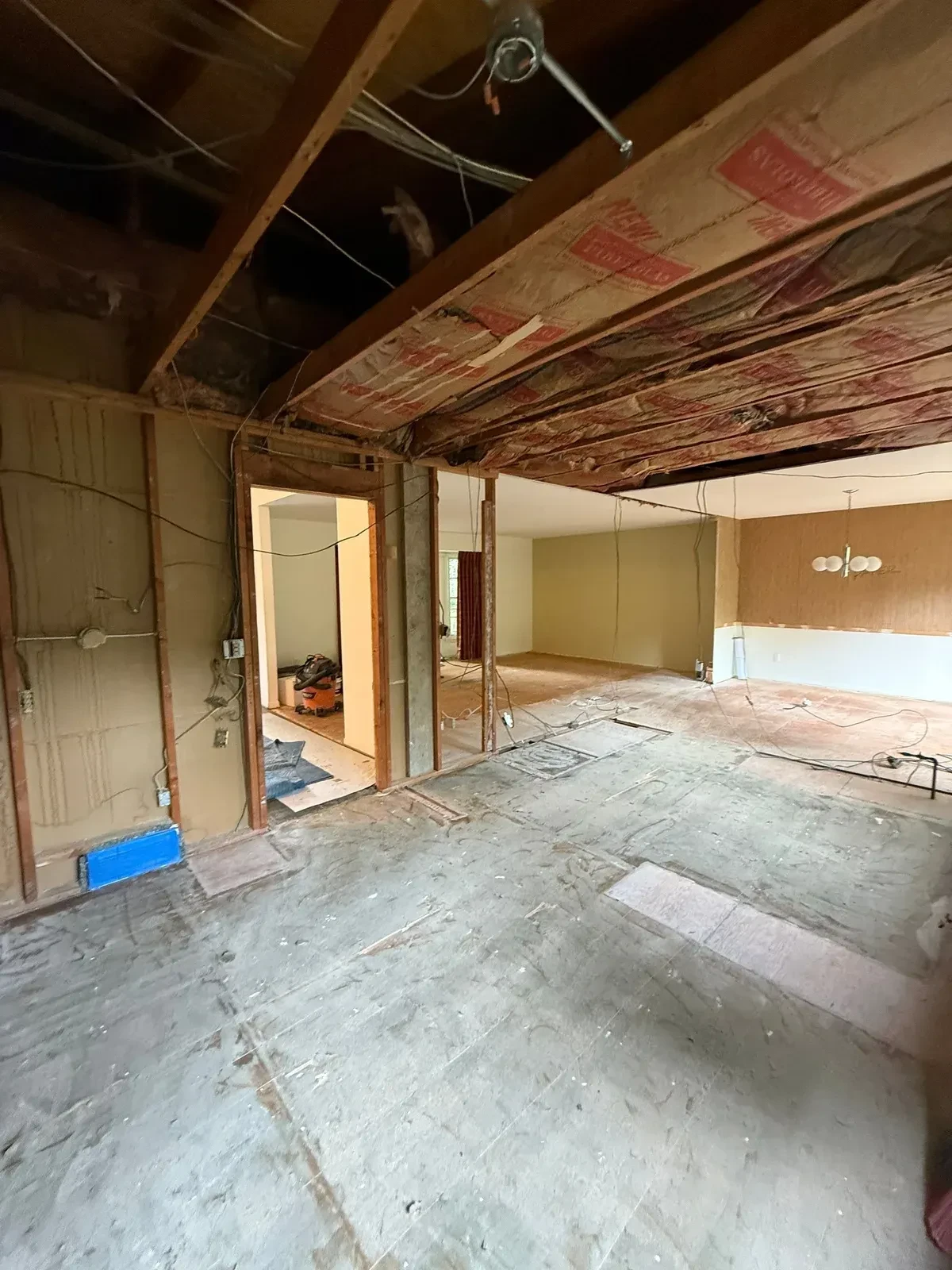 Gutted kitchen opened to the adjacent room with exposed wall framing, ceiling joists, insulation and stripped floor