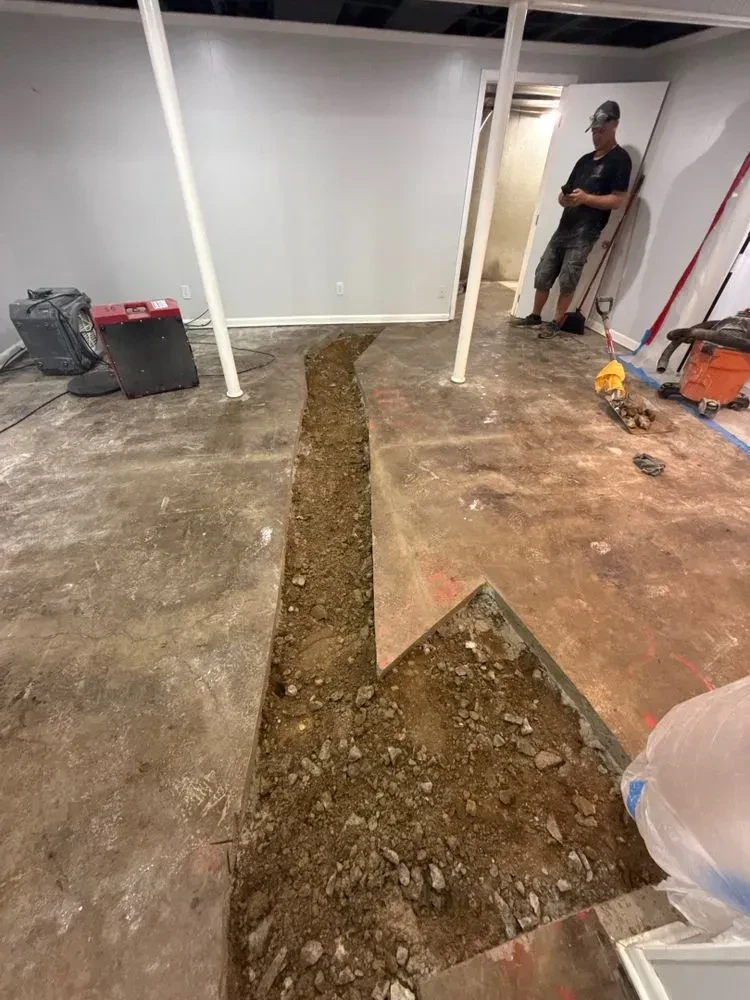 Long narrow basement concrete trench opened through the floor, with broken concrete and dirt visible