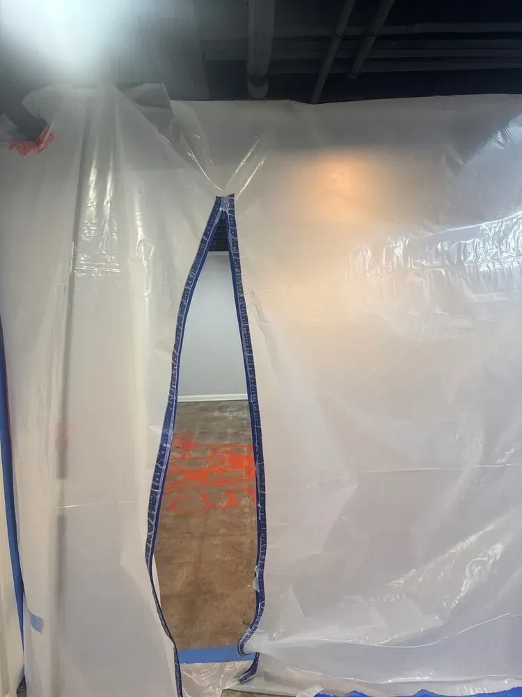 Close view through a zippered plastic containment opening into a basement room with orange floor markings