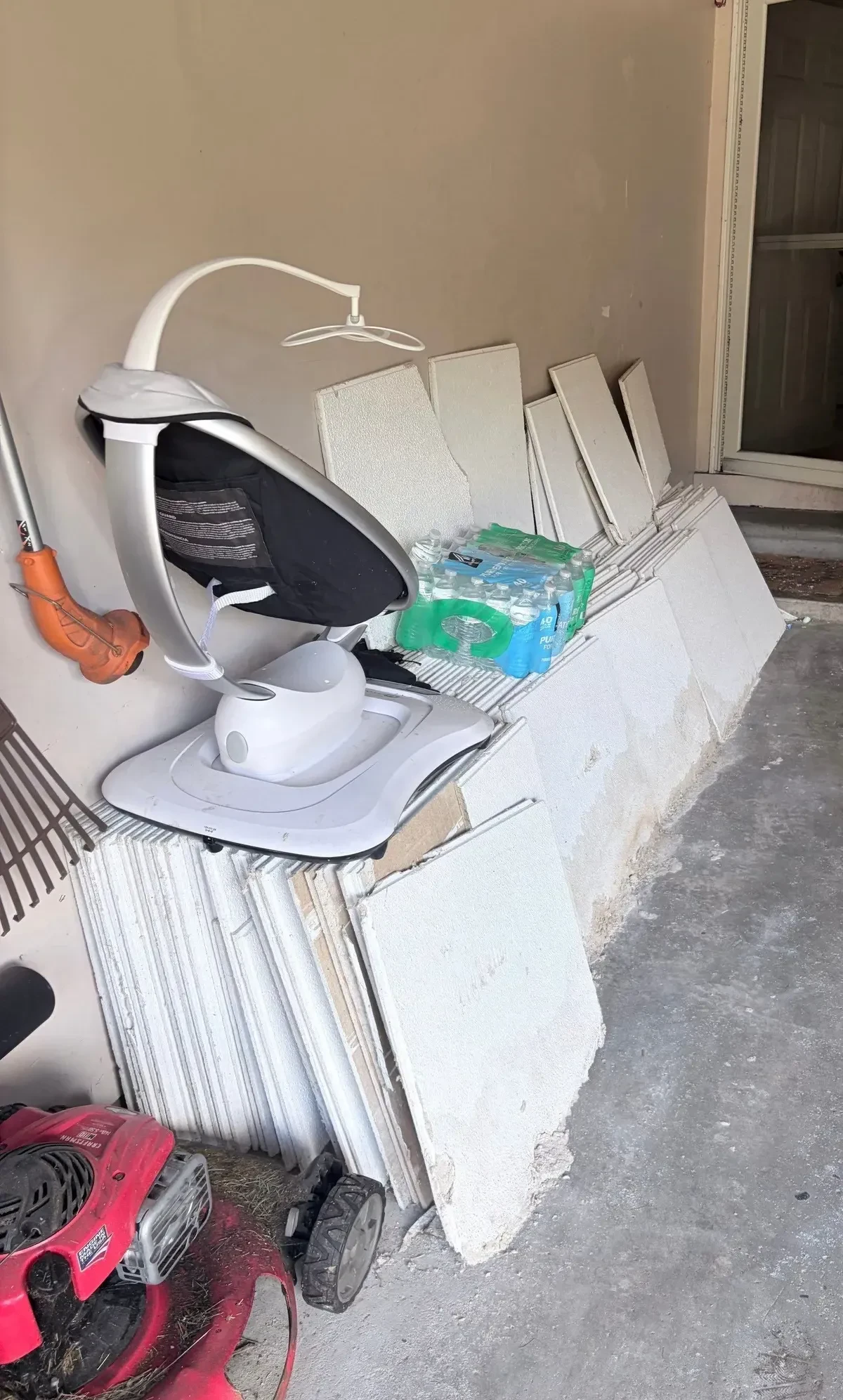 Basement ceiling panels stacked and leaning along a garage wall before removal, with a baby swing, bottled water and other staged items nearby