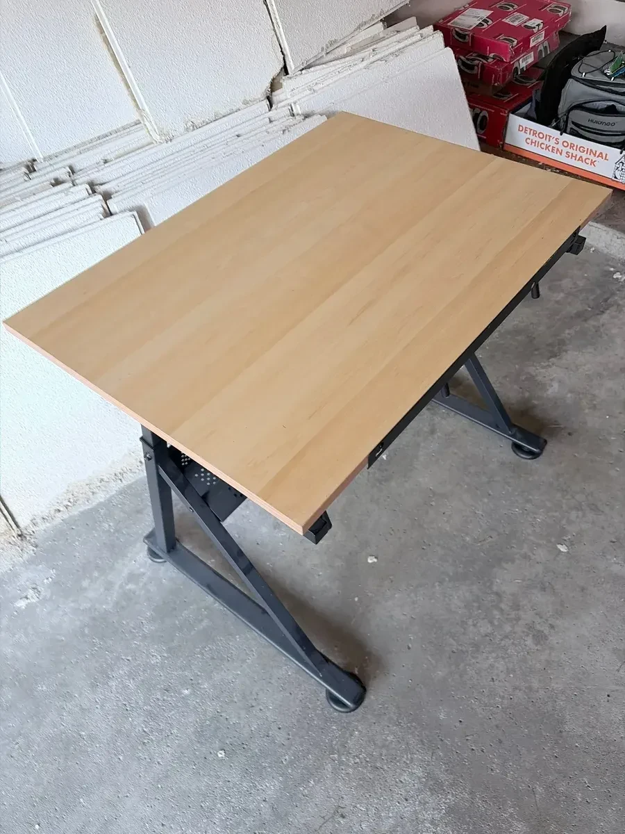 Wooden-top folding desk with black folding legs inside a garage, with white ceiling panels stacked behind it