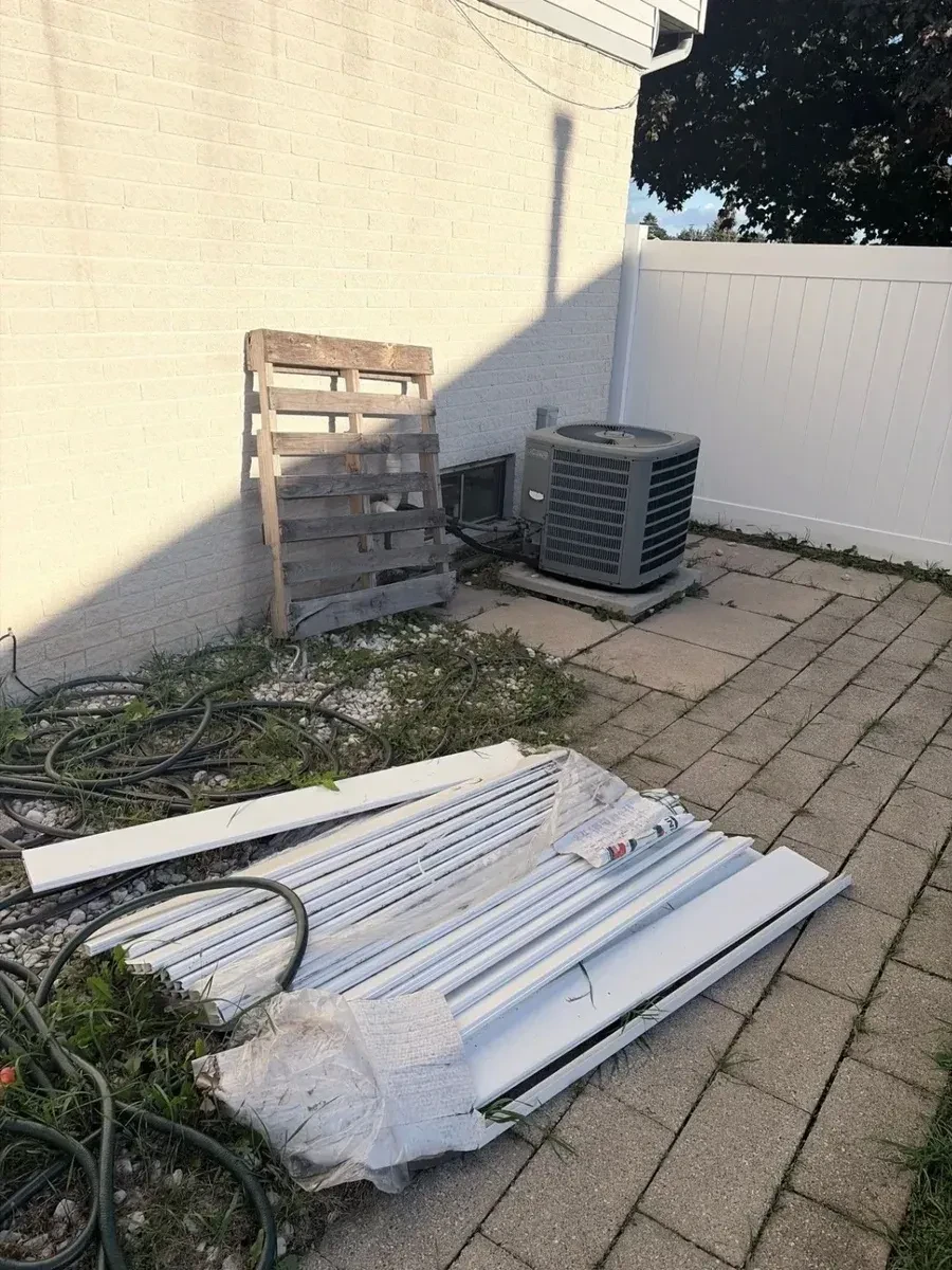 Wooden pallet, bundled leftover vinyl fence panels and coiled garden hoses beside a brick wall and AC unit on a paver patio in Clinton Township
