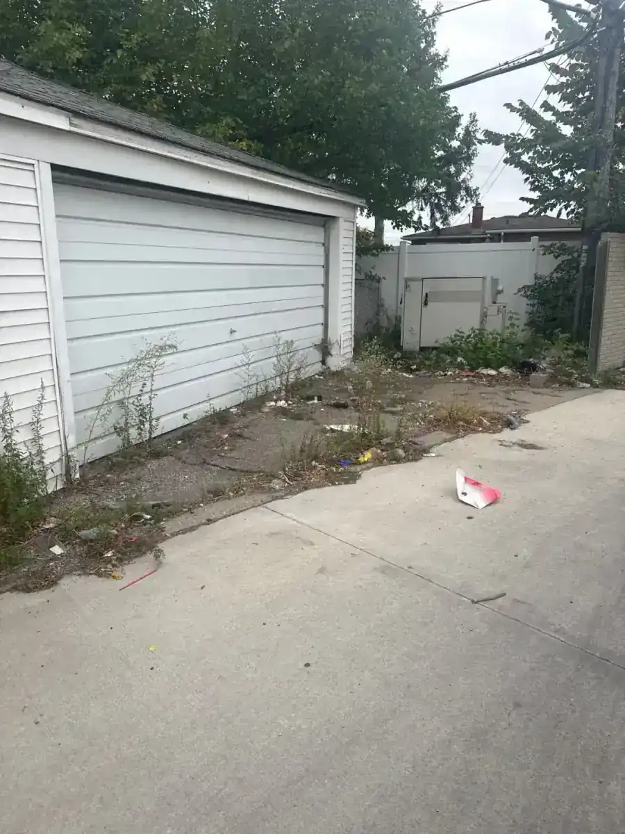 Litter and weeds along the garage wall and paved walkway before cleanup