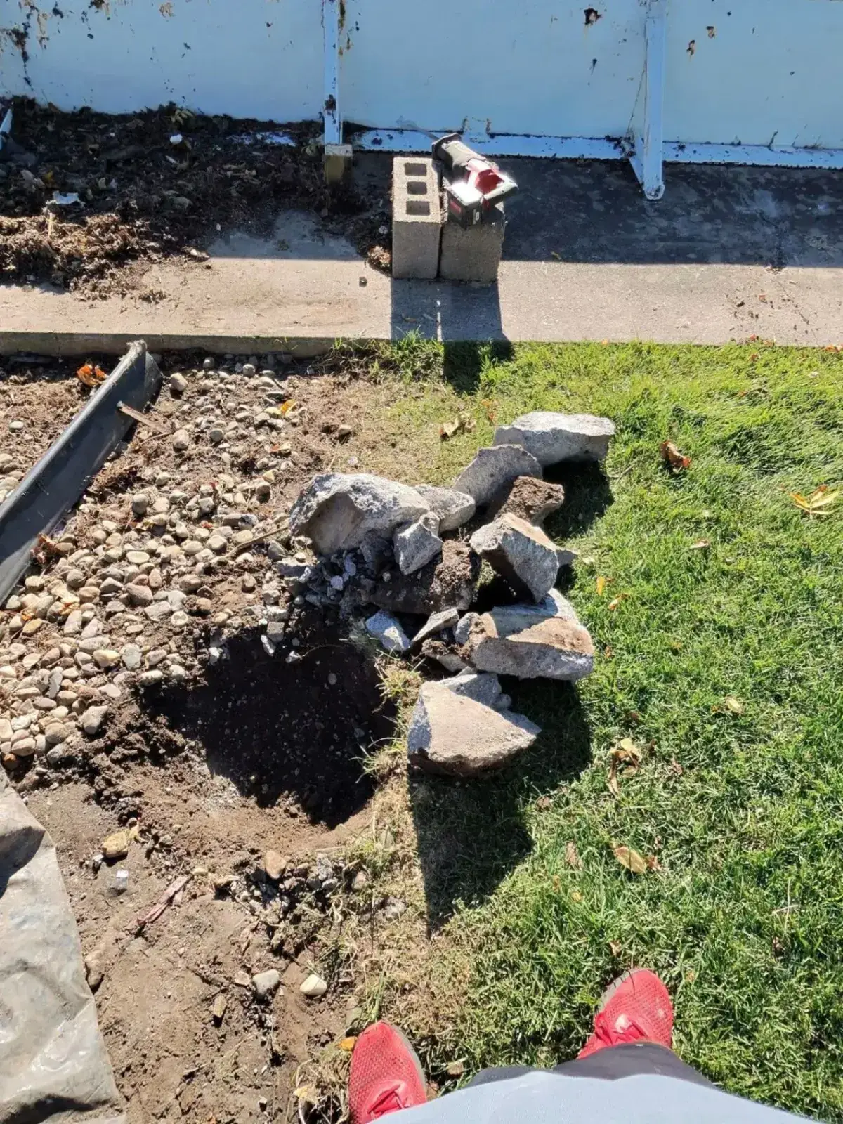 Broken concrete footing chunks beside the exposed hole where a waterfront deck post was set, with gravel and grass around it