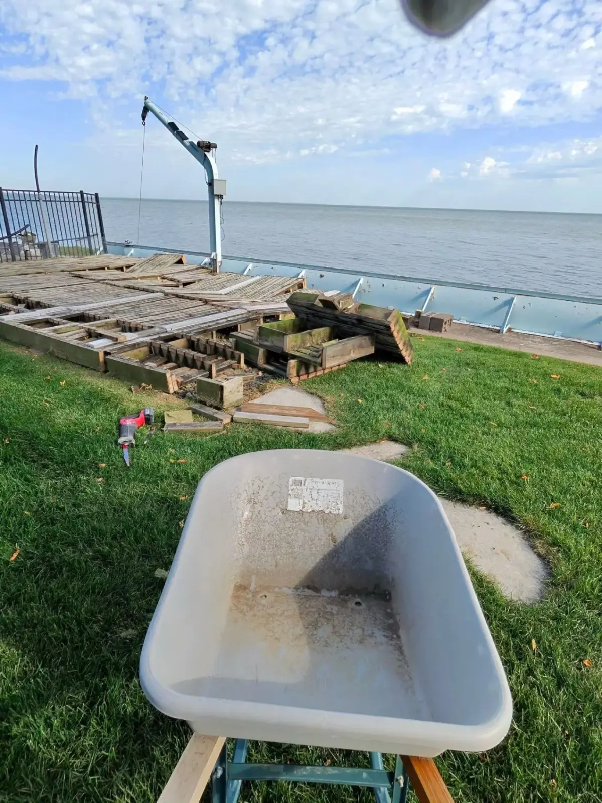 Waterfront deck demolition in progress with boards pulled up and framing exposed, a wheelbarrow in the foreground and a saw on the grass