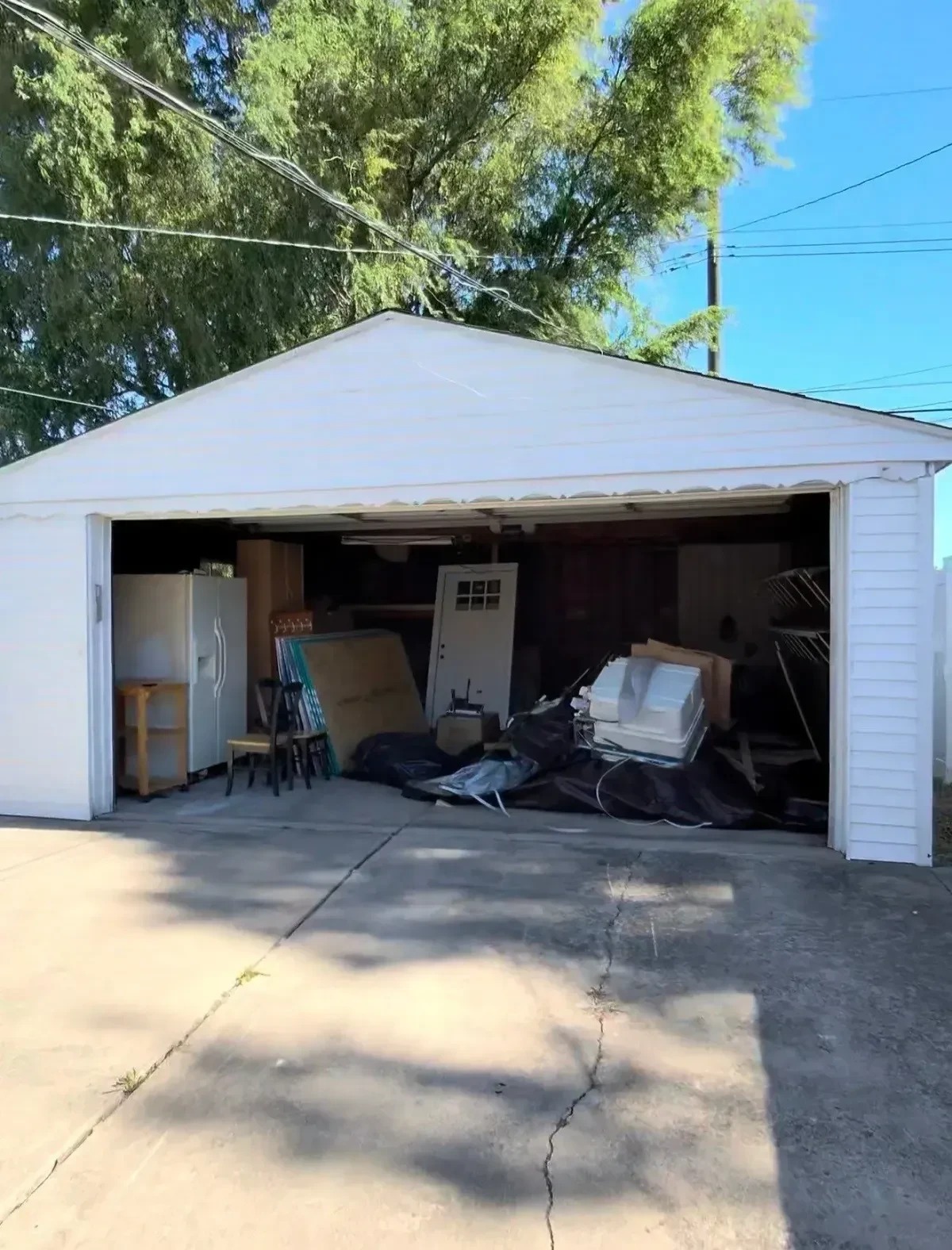 Open detached garage in St. Clair Shores before cleanout, with a refrigerator, loose panels, furniture and other debris visible inside