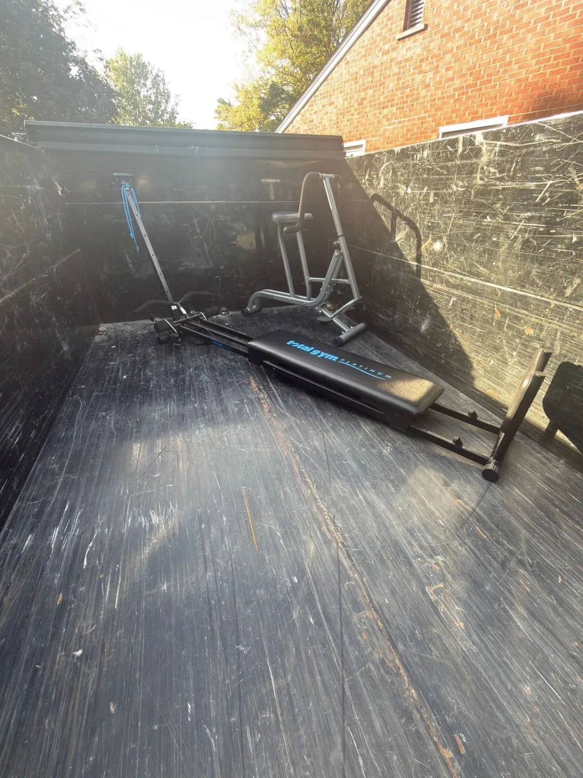 Total Gym and fitness exercise bike loaded inside a black Demohaulic dump trailer beside a brick house